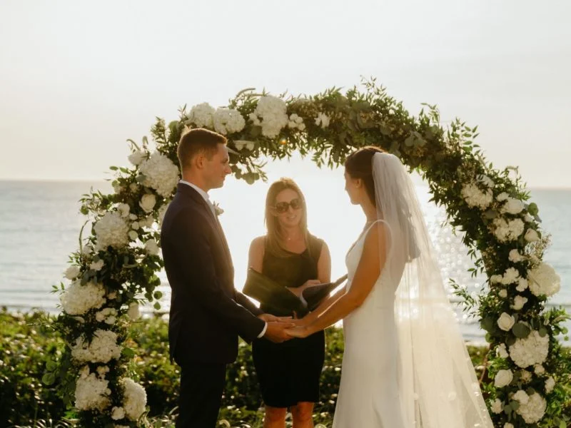 wedding ceremony with bride, groom, and officiant under white flower arch